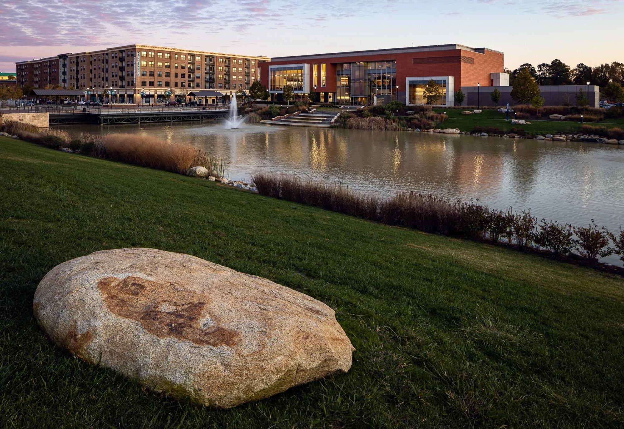 A boulder on the lawn beside the lake at Libbie Mill, with buildings beyond at dusk